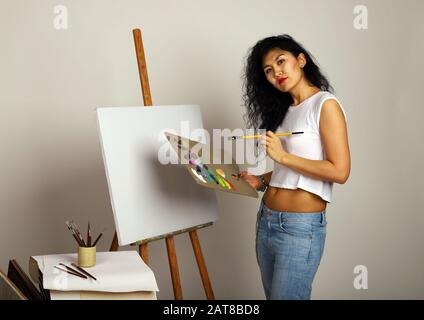 Der Künstler der Staffelei malt ein Bild. Attraktives Mädchen mit Pinselfarben. Eine leere Leinwand für ein neues Gemälde und ein Künstler mit Pinsel und Farben Stockfoto