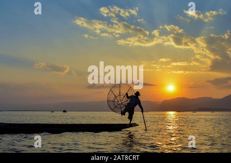 Traditioneller Fischer, der auf einem Bein auf einem Boot balanciert, einen Angelkorb hält, auf dem Inle-See bei Sonnenuntergang, Myanmar, angelt. Stockfoto