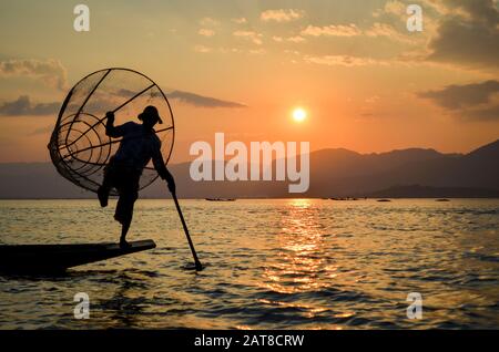 Traditioneller Fischer, der auf einem Bein auf einem Boot balanciert, einen Angelkorb hält, auf dem Inle-See bei Sonnenuntergang, Myanmar, angelt. Stockfoto