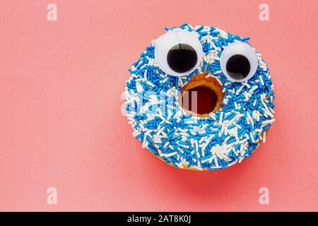 Vanille Dip Donut mit schwarzen Wiggle Googly Augballs auf einem Farbigen blau-weißen Besprengung auf einem rosafarbenen Hintergrund. Stockfoto