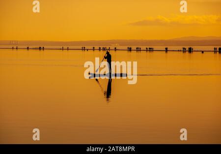 Man steht beim Sonnenuntergang auf dem Paddel-Boarding in Silhouette, auf dem Bootssee Marine Lake West Kirby auf der Wirral mit der Dee-Flussmünde im Hintergrund Stockfoto