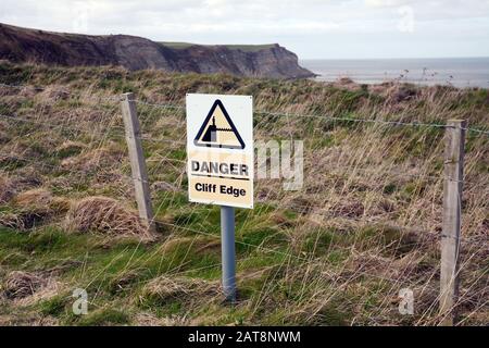 Ein Gefahrenschild, das vor einer Felskante warnt, entlang des Cleveland Way, einem Wanderweg im North York Moors National Park, Yorkshire, England, Großbritannien. Stockfoto