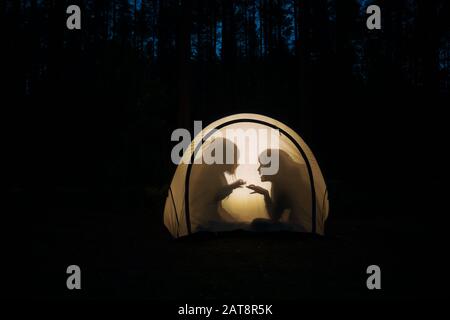 Silhouetten von Kindern, die nachts im Zelt spielen, machen Schattenpuppen mit Taschenlampe und genießen Sommerferien Stockfoto