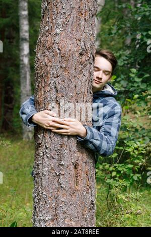 Teenager-Junge umarmende Pinie in einem Sommerwald, der sich an Baumstamm lehnt, mit geschlossenen Augen, die die Natur umarmen Stockfoto