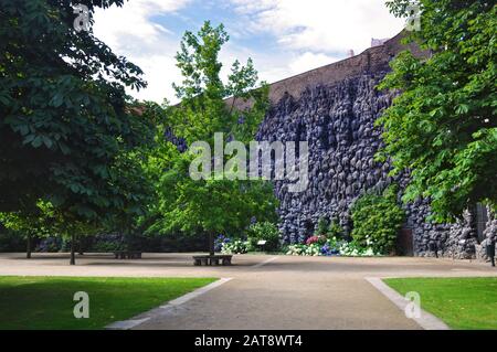 Blick auf die leere Gasse, die zum Wallensteingarten führt, seine graue Tropfsteinmauer mit Felsformationen, grünen Bäumen und Blumen unter blauem Himmel und weißem Clou Stockfoto