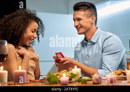 Romantisches Datum. Junges multiethnisches Paar zu Hause, das einen Mann zum Abendessen hat, der der Frau vorschlägt, mit einem lächelnden Proposal Stockfoto