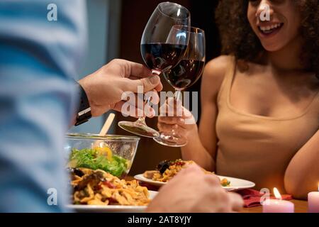 Romantisches Datum. Junges multiethnisches Paar zu Hause sitzen am Tisch Essen Abendessen jubelt mit Gläsern Wein und lacht freudig nah Stockfoto