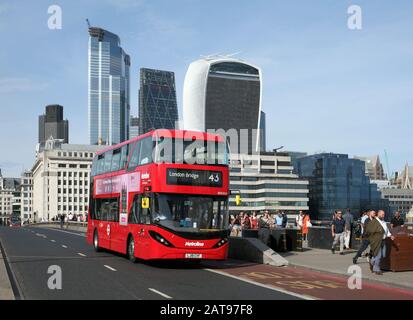 Ein elektrischer (batteriebetriebener) Doppeldeckerbus, der die London Bridge, London, überquert. Hintergrund: Hochhäuser in der City of London. Stockfoto