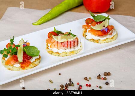 Runde Sandwiches mit Lachs und rotem Kaviar. Weiß die lange quadratische Platte liegt auf einem weißen und grauen Wickelpapier, überlagert mit Pfeffererbsen, Tomaten, Stockfoto