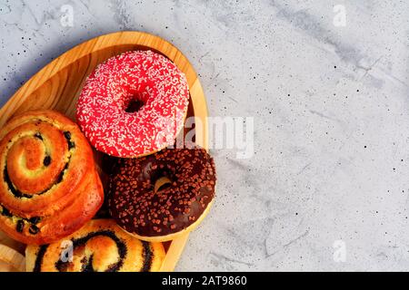 Köstliche Schokoladendonuts und Brötchen mit Mohnsamen und Rosinen liegen auf einem ovalen Holztablett und einer grauen Betonoberfläche. Stockfoto