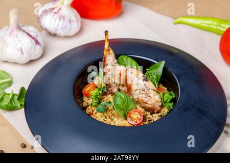 Gebratenes Kaninchenbein mit Reis auf einem runden schwarzen Teller. Garnierung ist mit Grüns und Tomaten dekoriert. Platte auf einem Hintergrund aus grauem und weißem Wickelpap Stockfoto