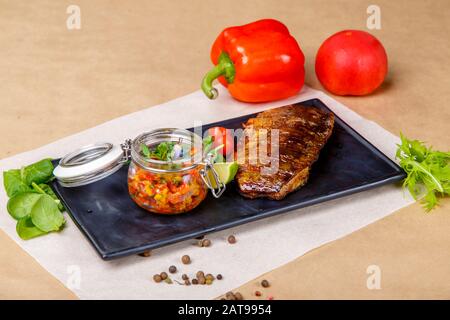 Ein saftiges großes Stück gegrillten Fisches sitzt auf einem quadratischen schwarzen Teller. Neben einem Krug mit spezieller Soße, einer Scheibe Kalk, Spinat, Tomaten. Die Platte ist eingeschaltet Stockfoto