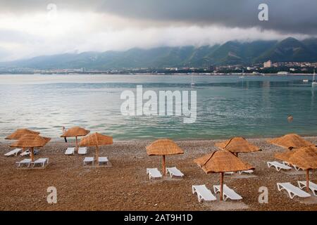 Abend in der Gelendzhik Bay. Im Vordergrund steht ein Kieselstrand, Sonnenliegen und Strohschirme aus der Sonne. In der Bucht auf dem Parkplatz des YA Stockfoto