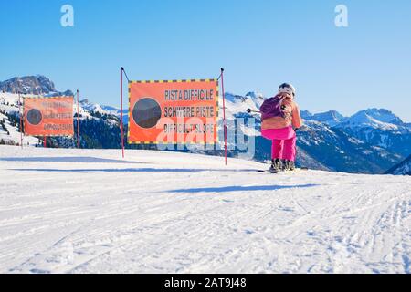Frau Skifahrerin, die eine schwarze Skipiste startet, die nur Experten vorbehalten ist und zwei Warnschilder übergibt, die Eine Schwierige Piste auf Italienisch, Deutsch und Englisch sagen, dur Stockfoto
