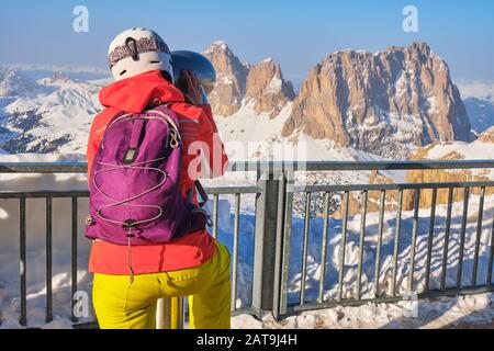 Frau Skifahrerin, die einen Helm trägt und durch Münzferngläser in Richtung Sassolungo (Saslonch, Langkofel) blickt, Gipfel in den Dolmen, Italien, auf der sonne Stockfoto