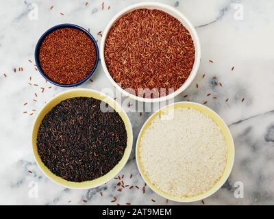 Keramikschüsseln mit einer Vielzahl von Getreide und Reis, einschließlich rotem Quinoa, rotem Reis, schwarzem Reis und weißem Reis auf einer Marmorplatte. Stockfoto