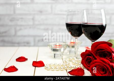 Valentinstag Konzept. Zwei Gläser Wein, rote Rosen, Perlenschnur, Rosenblätter und brennende Kerzen auf einem weißen Holztisch mit Platz für Kopien Stockfoto