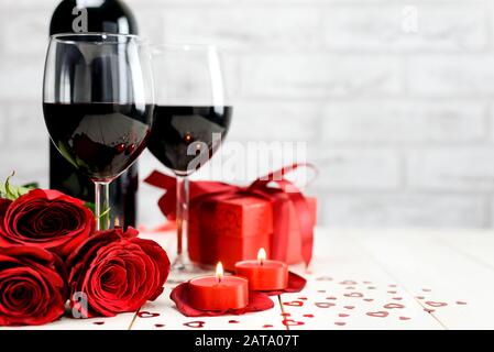 Valentines Tageskonzept. Zwei Gläser Wein, rote Rosen, Weinflasche, Geschenkbox und brennende Kerzen auf einem weißen Holztisch mit Platz für Text. Se Stockfoto