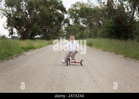 Ein kaukasischer australischer Junge, der auf einem roten Dreirad auf einer unbefestigten Straße im Land fährt Stockfoto