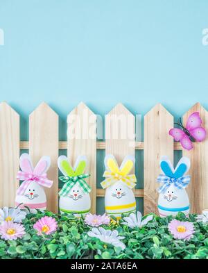 Mehrere lustige Osterhasen mit bunten Schleifen aus Ostereiern, umgeben von Gras, Blumen und Schmetterlingen, gegen den blauen Himmel, mit Platz Stockfoto