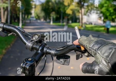 Eine Zigarette in der Hand eines rauchen Radfahrer, Fahrten mit dem Fahrrad auf dem Hintergrund der asphaltierte Weg des City Park Stockfoto