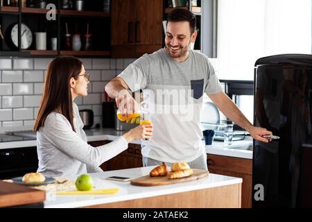 Ein Paar in der Küche essen Frühstück mit Orangensaft und Gebäck am Tisch und er öffnet einen Kühlschrank Stockfoto