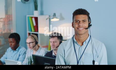 Portrait des multirassischen Kundendienst-Bedieners im Headset, das an die Kamera lächelt. Arbeitskollegen stehen im Hintergrund. Stockfoto
