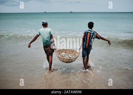 Zwei srilankische Männer, die Fische in einem Bambo-Korb am Trincomalee Strand tragen und im Ozean waschen. Stockfoto