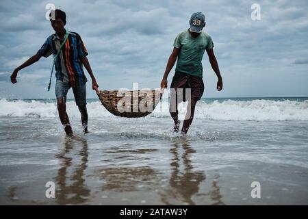 Zwei srilankische Männer, die Fische in einem Bambo-Korb am Trincomalee Strand tragen und im Ozean waschen. Stockfoto