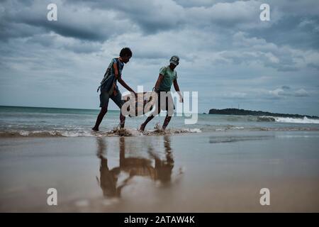 Zwei srilankische Männer, die Fische in einem Bambo-Korb am Trincomalee Strand tragen und im Ozean waschen. Stockfoto