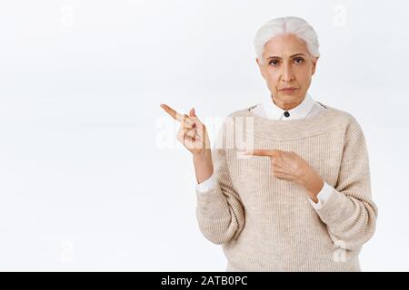 Wütende, seriös aussehende und streng elegant arbeitende Senior-Dame mit grauem gekämmtem Haar, Stirnrunzeln, die die Erklärungen der Nachfrage schmunzeln und die Finger nach links zeigen Stockfoto