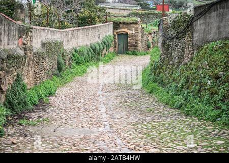 Ländliche gepflasterte und nasse Straße zwischen übereinanderliegenden Steinmauern und traditioneller Bauweise, in regnerischer Umgebung, in Serradilla (Cáceres), wes Stockfoto