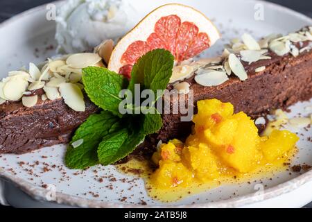 Brownie Schokoladendessert mit einer Schaufel weißer Eiscreme, Scheiben Mango und einer Scheibe Grapefruit. Im Hintergrund steht ein Cappuccino. Auf einem dunklen Bac Stockfoto