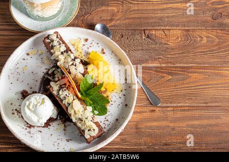 Brownie Schokoladendessert mit einer Schaufel weißer Eiscreme, Scheiben Mango und einer Scheibe Grapefruit. Im Hintergrund steht ein Cappuccino. Auf einem Holzn Stockfoto