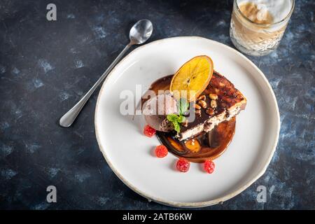 Schokoladendessert mit einer Schaufel Kaffeeissahne, Scheiben getrockneter Kirsche und einer Scheibe Orange. Garniert mit Minzblättern. Cappuccino im Backgr Stockfoto