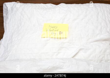 Abschluss einer gelben kleinen Notiz mit dem Text Happy Valentines Day darin geschrieben, auf dem Kissen weiße Bettlaken Stockfoto