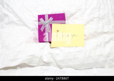 Nahaufnahme einer kleinen gelben Notiz mit handschriftlicher Schrift ich liebe dich auf weißen Bettlaken und einem Geschenkkasten, Valentine-Konzept Stockfoto