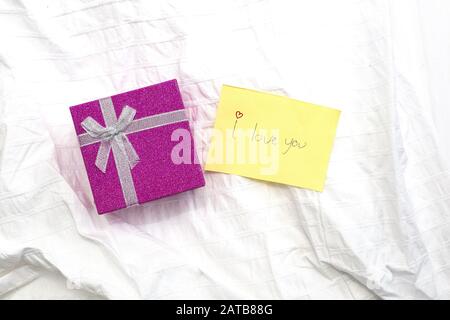 Nahaufnahme einer kleinen gelben Notiz mit handschriftlicher Schrift ich liebe dich auf weißen Bettlaken und einem Geschenkkasten, Valentine-Konzept Stockfoto