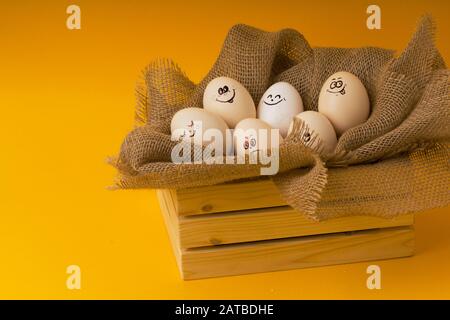 Rustikaler Korb mit lustigen Eiern mit gemalten Gesichtern, die unterschiedliche Emotionen auf gelbem Hintergrund ausdrücken. Nahaufnahme . Stockfoto