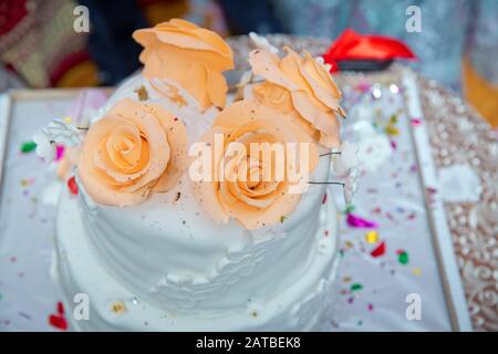 Weißer Kuchen gelb vier Blumen. Beige 4 gestufte Hochzeitstorte mit zarten Rosen stehen auf dem Tisch auf Stoffhintergrund. Stockfoto