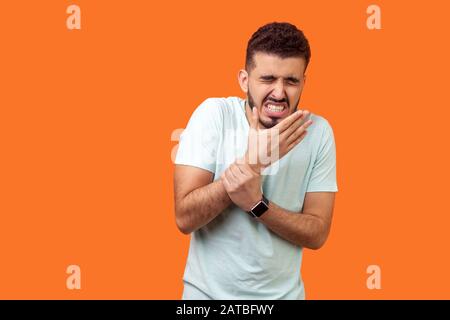 Verletzter Arm. Porträt des brünetten Mannes mit Bart in zwanglosem weißen T-Shirt mit schmerzhaftem Handgelenk, mit Handstauchung oder Arthritisentzündung. Indo Stockfoto