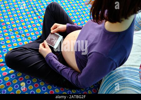 Ein junges, schwangeres kaukasisches Mädchen betrachtet den Röntgenstrahl ihres Babys und sitzt nachdenklich auf dem Bett. Stockfoto
