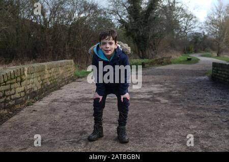 Ein 8-jähriger Junge beugt sich mit einem schlammigen Gesicht und Stiefeln über die Knie und trägt einen Parka-Mantel mit Blick auf die Kamera, Winter 2020 Stockfoto