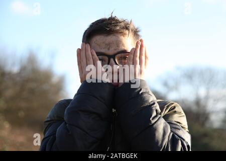 Ein Junge, der eine Brille im Alter von 12 Jahren trägt, bedeckt sein schlammiges Gesicht mit den Händen und zeigt die Kamera Stockfoto