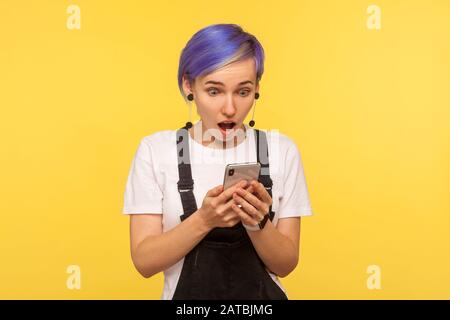 Porträt eines staunenden Hüftmädchen mit violettem Haar in Overalls mittels Handy, SMS oder Nachrichten mit schockierenden Nachrichten lesen, überrascht betrachtet Stockfoto