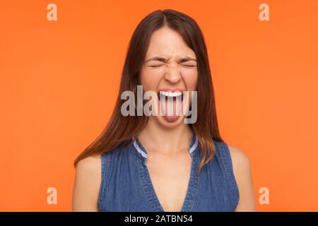 Ungehorsam verrückte brünette Frau in Denimkleidung, die mit geschlossenen Augen Zunge herausstreckt und grimacing, klaffend mit derben, frechen Ausdrücken zeigt. Indo Stockfoto