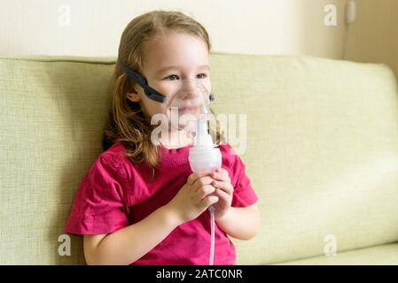 Kleines Mädchen hält Inhalationsmaske zu Hause. Krankes Kind atmet durch einen Vernebler. Baby mit Geräten zur Behandlung von Asthma oder Bronchitis. Konzept der Behandlung Stockfoto