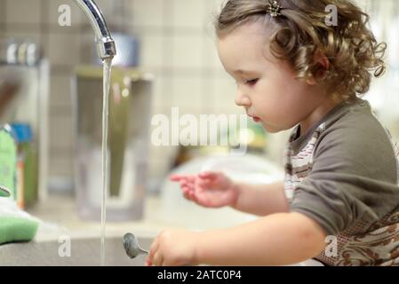 Hübsches Kind wäscht Geschirr in der Küche. Nettes Kleinkind lernt den Haushalt. Wunderschönes zweijähriges Mädchen wäscht Löffel im Waschbecken. Adorable Kind houses cl Stockfoto