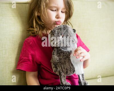 Kleines Mädchen hält Inhalationsmaske zu Hause. Lustige Kinder spielen mit Spielzeug und Vernebler. Baby mit Geräten zur Behandlung von Asthma oder Bronchitis. Konzept der Behandlung Stockfoto