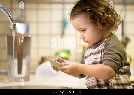 Ein süßes zweijähriges Mädchen wäscht in der Küche Geschirr Stockfoto